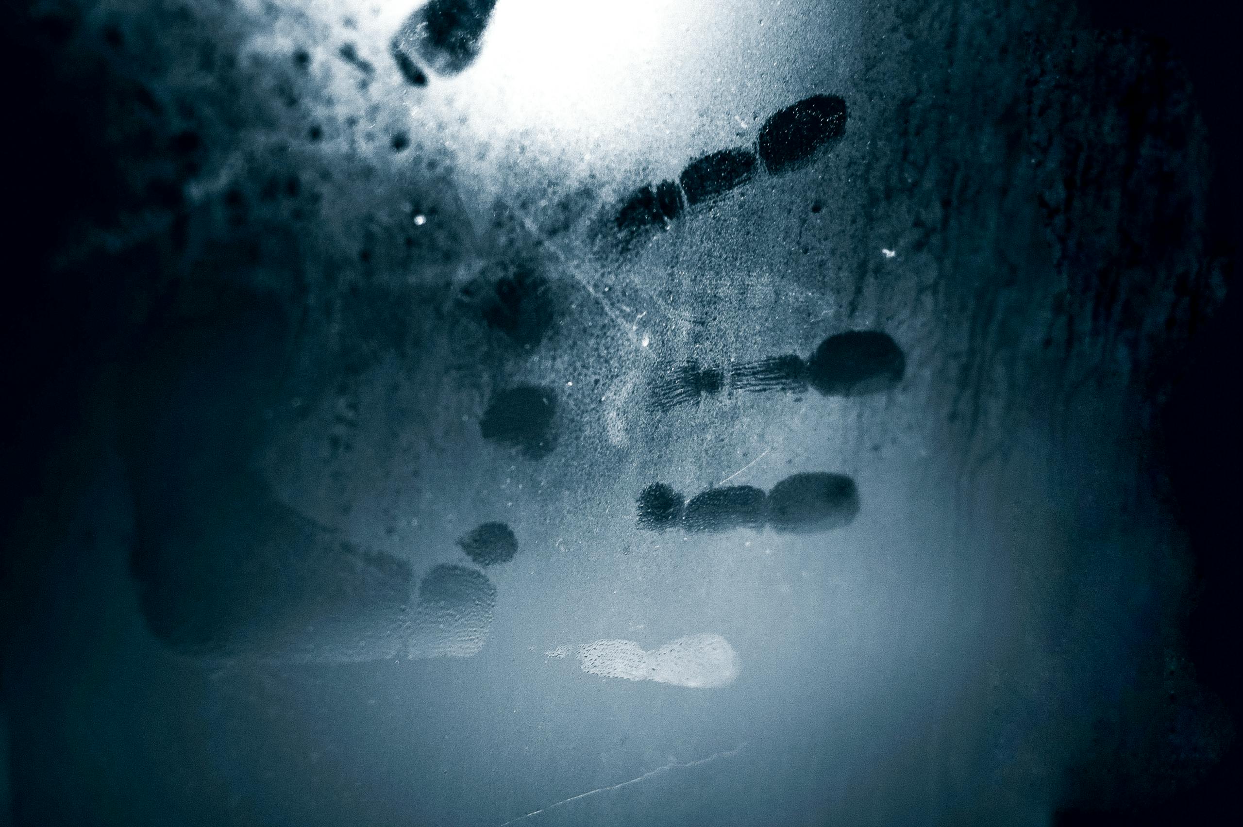 A mysterious handprint on a fogged glass surface illuminated by eerie blue light creates a dark, moody atmosphere.